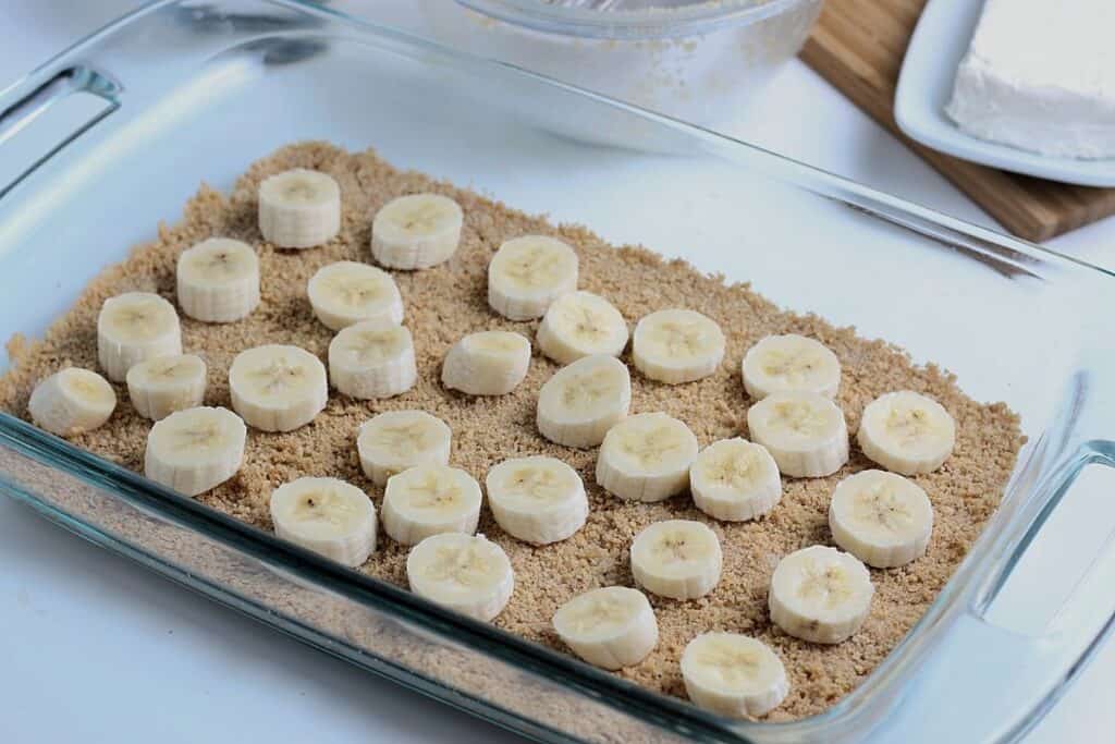 Graham cracker crust layered in bakeware with banana slices on top.