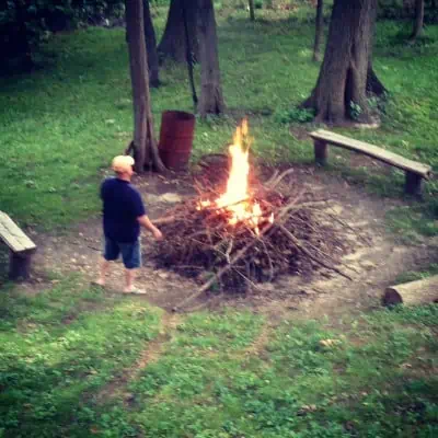 A man standing by a large pile of sticks and a bonfire. 