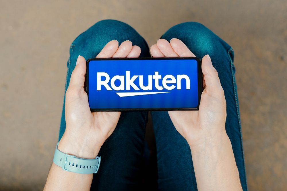 A person holds a smartphone showing the Rakuten logo, resting on their lap with both hands visible.