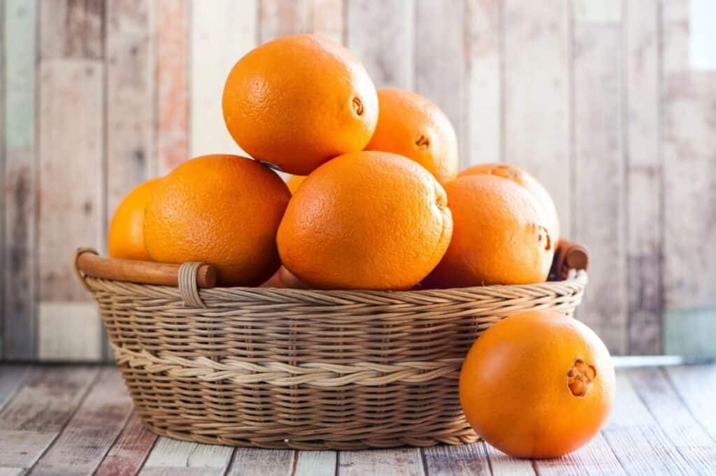 A wicker basket filled with whole oranges sits on a wooden surface, with one orange resting beside the basket; wood panel wall in the background—perfect for exploring creative ways to use orange peels.