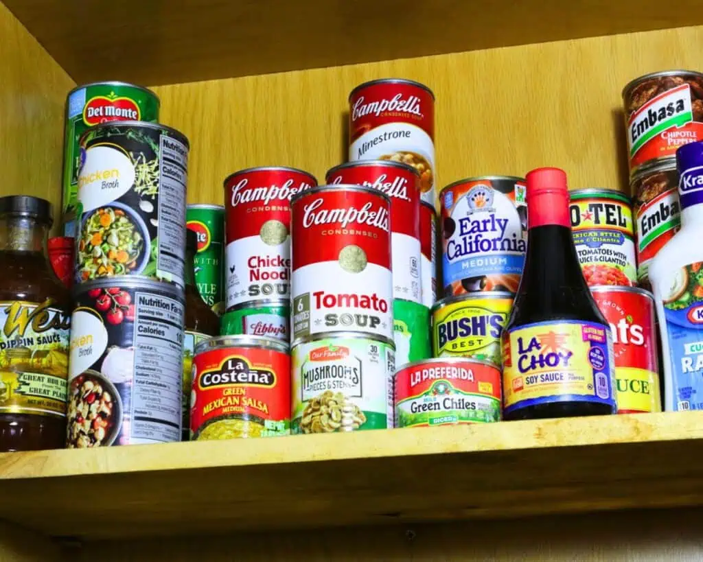 A wooden pantry shelf showcases how to build a stockpile, brimming with various canned goods and sauces, including soups, green chiles, olives, and soy sauce.