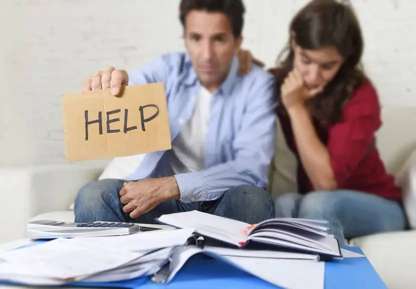 A couple with their budget and financial books open. The husband is holding up a \"help\" sign.