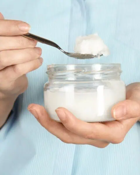 A person holds a glass jar of coconut oil in one hand and scoops some out with a spoon using the other, exploring different ways to use coconut oil in their daily routine.