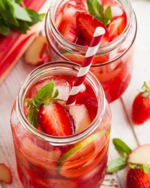 Two mason jars of pink strawberry rhubarb lemonade with ice, fresh strawberries, and striped straws. Rhubarb and strawberry garnish surround the drinks on a wooden surface.