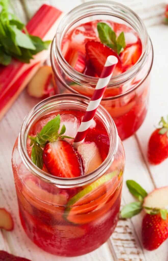 Two mason jars of pink strawberry rhubarb lemonade with ice, fresh strawberries, and striped straws. Rhubarb and strawberry garnish surround the drinks on a wooden surface.