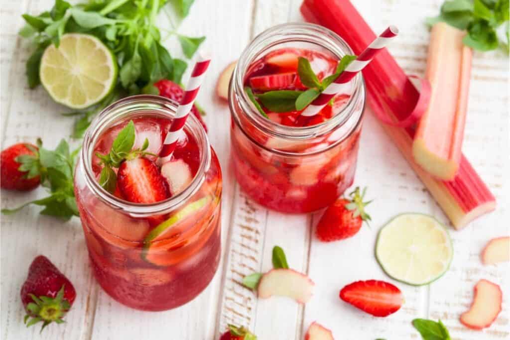Two glass jars of refreshing strawberry rhubarb lemonade with striped straws sit on a white wooden table, surrounded by fresh strawberries, rhubarb, lime slices, and vibrant green leaves.