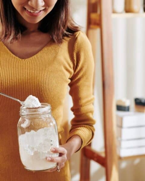 A woman in a mustard sweater scoops white powder from a large glass jar with a spoon in a bright kitchen, exploring new uses for baking soda.