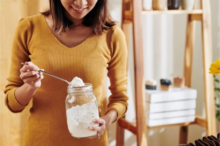 A woman in a mustard sweater scoops white powder from a large glass jar with a spoon in a bright kitchen, exploring new uses for baking soda.