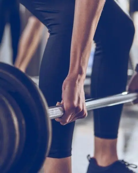Person gripping a loaded barbell, preparing to lift weights in a gym setting; focus is on hands and bar with blurred background—perfect for those aiming to cut gym costs by mastering lifts at home.