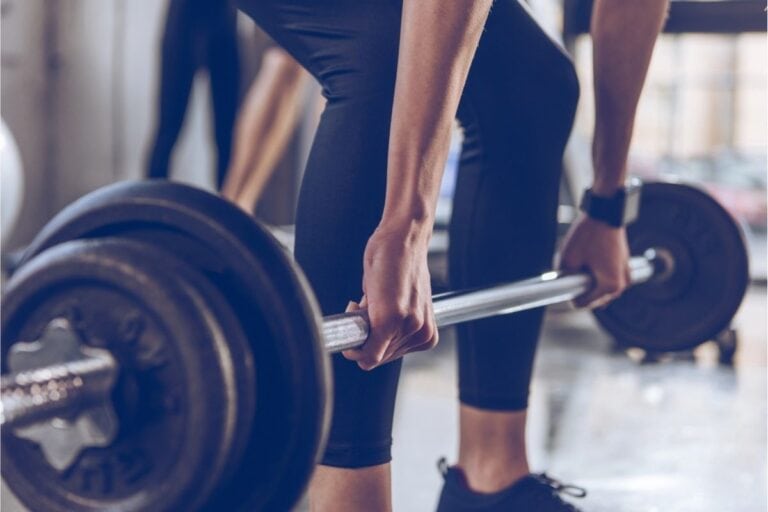 Person gripping a loaded barbell, preparing to lift weights in a gym setting; focus is on hands and bar with blurred background—perfect for those aiming to cut gym costs by mastering lifts at home.