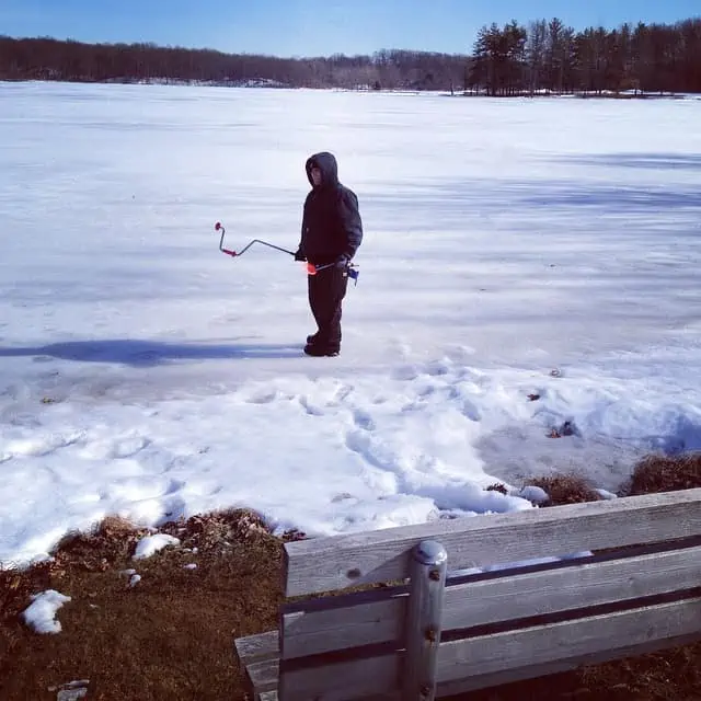 A man standing in the middle of a frozen lake going ice fishing.