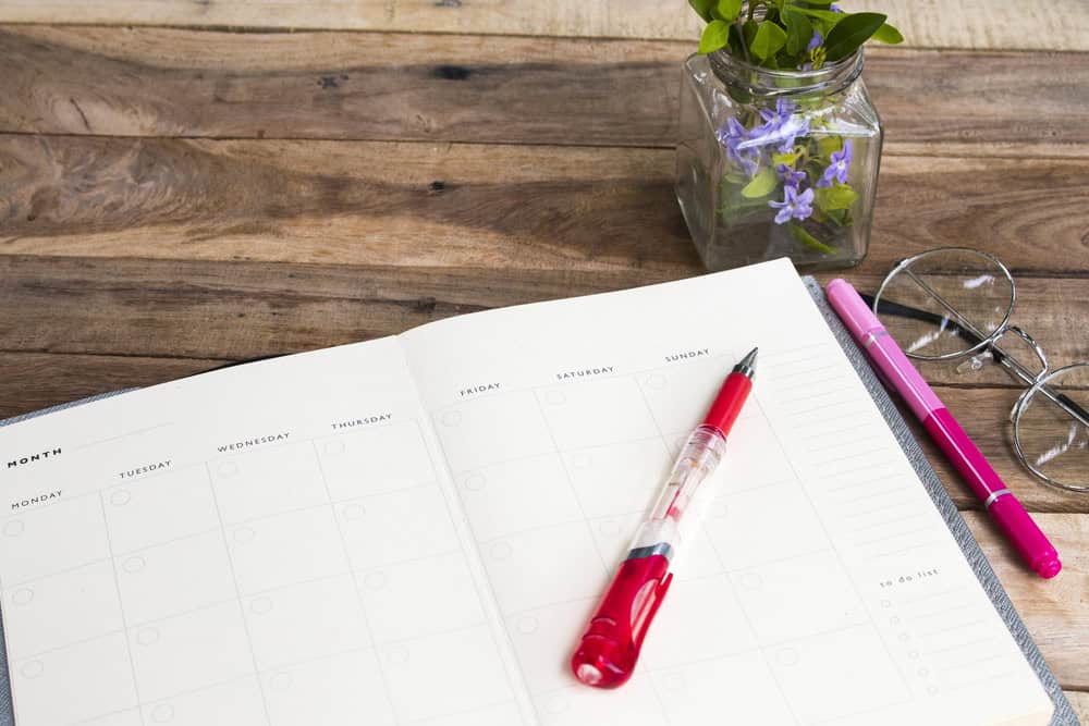 An open planner rests on a wooden table, flanked by two pens and eyeglasses, ready to jot down ideas on how to create a 30-day meal plan rotation. Nearby, a small vase holds purple flowers, adding a splash of color to the organized scene.