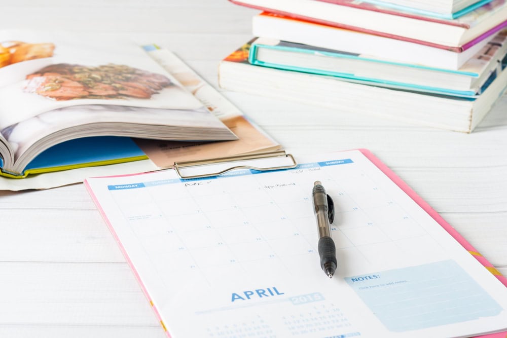 Calendar open to April with a pen on top, beside stacked books on a white wooden surface—perfect for jotting down how to create a 30-day meal plan rotation.