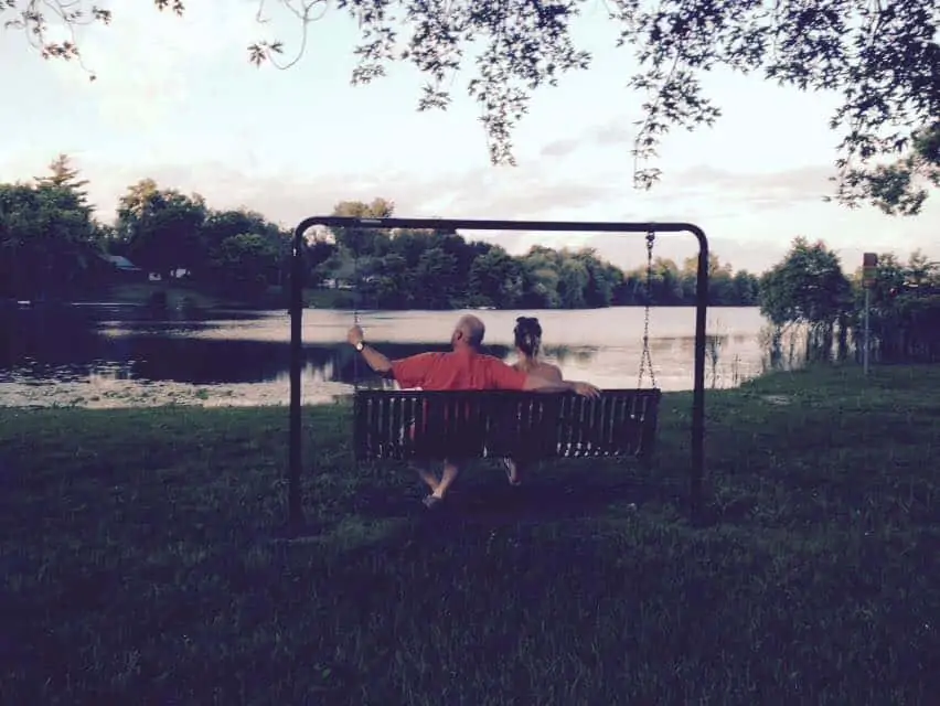 A man and woman sitting next to each other on a swinging bench by a body of water.