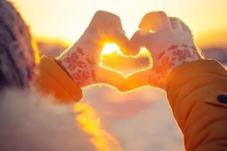 A woman holding her hands in a heart-shape against the snow and sunshine.
