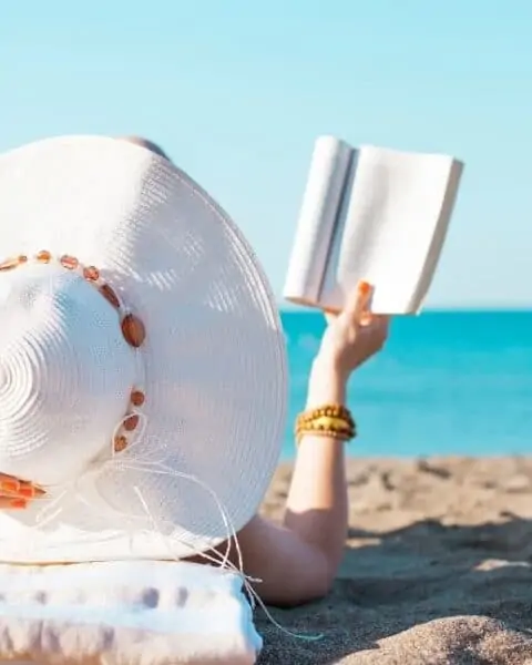 A woman in a sunhat reading a book on the beach.