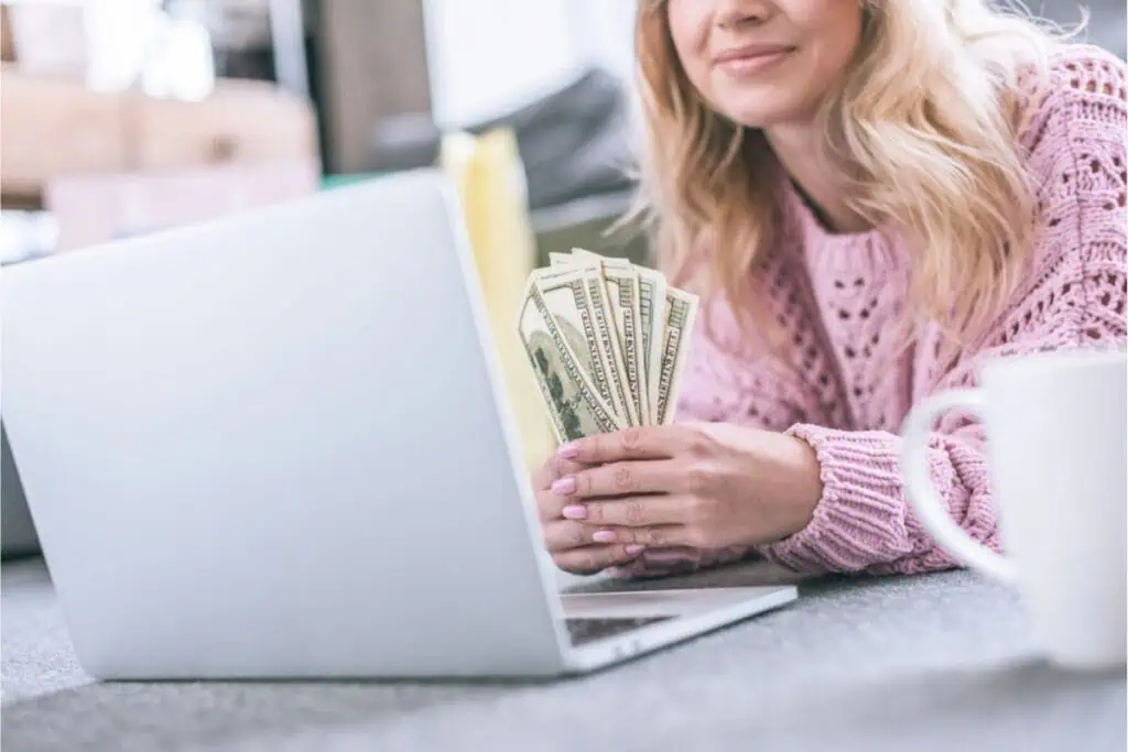 A person sits at a laptop, a stack of U.S. dollar bills in hand, contemplating how to make money selling stuff online, with a white mug nearby.