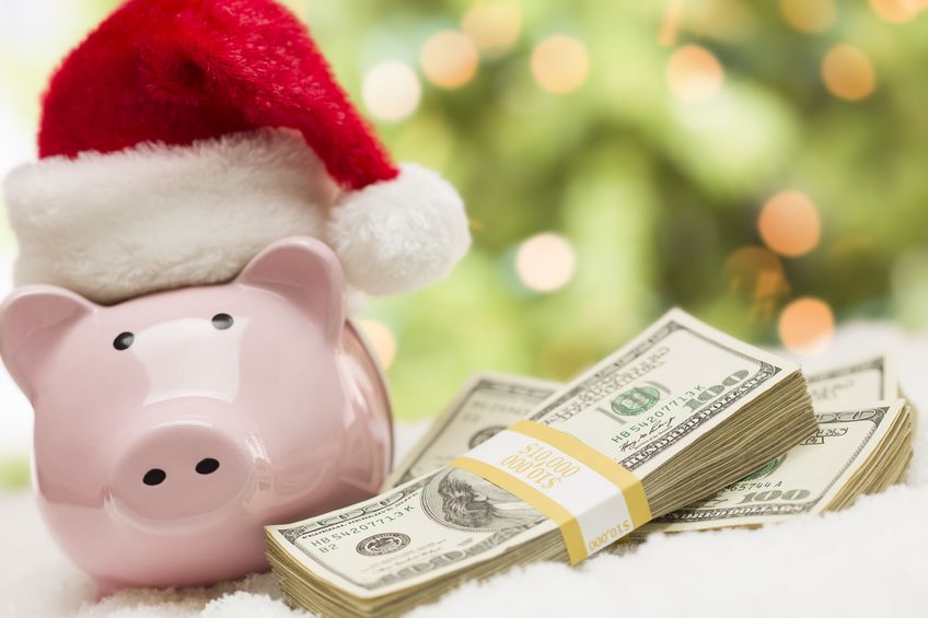 pink piggy bank wearing Santa hat near stacks of hundreds of dollars of money on snowflakes.