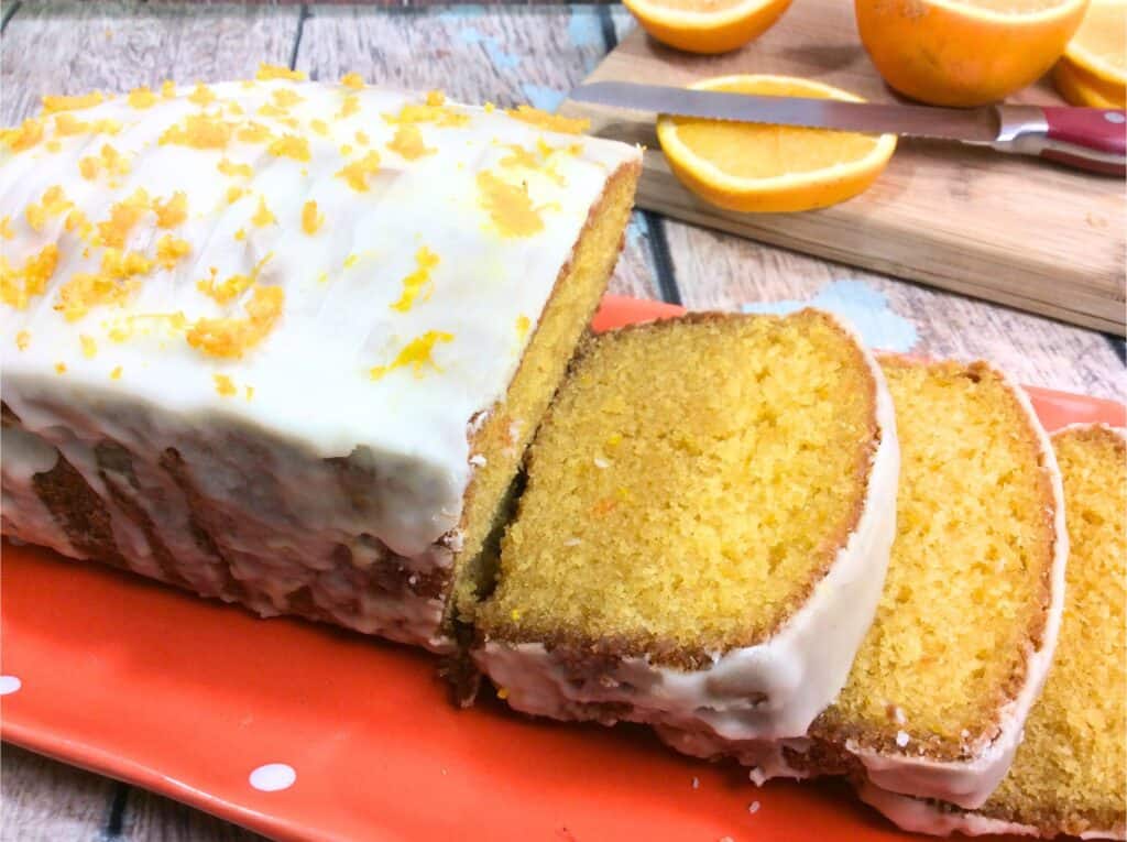 Partially sliced on a red platter, this orange cake with white icing and orange zest on top embodies the Best Orange Creamsicle Cake Recipe, complete with sliced oranges on a cutting board in the background.