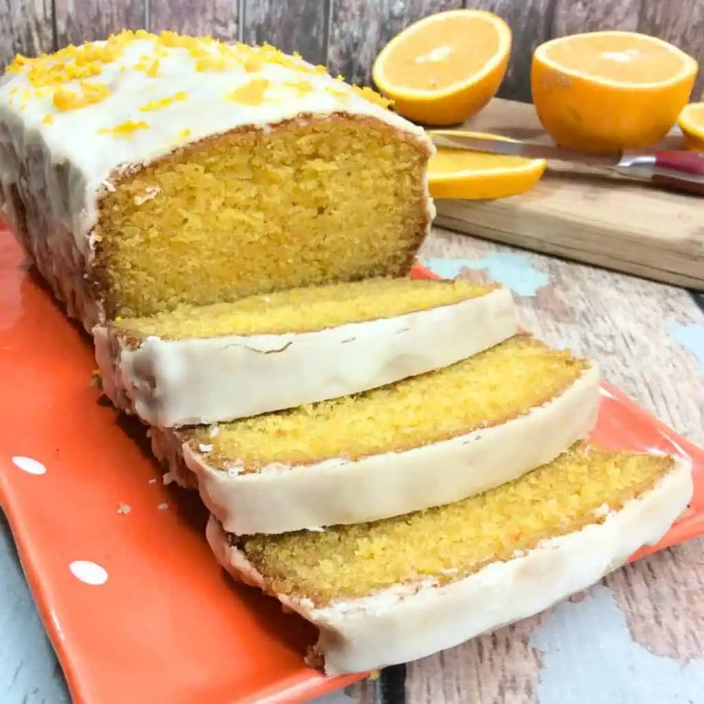 Sliced orange loaf cake with white icing on a red plate, garnished with orange zest, reminiscent of the best Orange Creamsicle Cake Recipe. Halved oranges and a knife linger in the background, adding to the vibrant display.