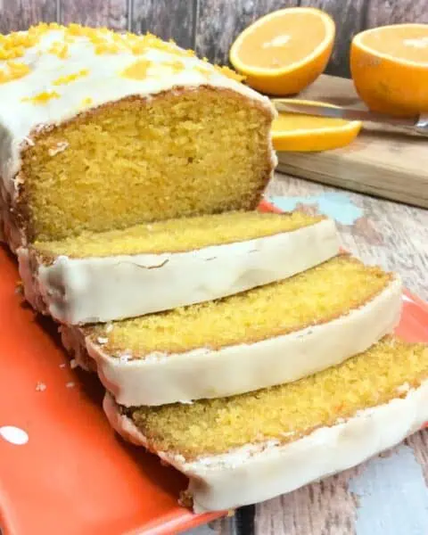 Sliced orange loaf cake with white icing on a red plate, garnished with orange zest, reminiscent of the best Orange Creamsicle Cake Recipe. Halved oranges and a knife linger in the background, adding to the vibrant display.