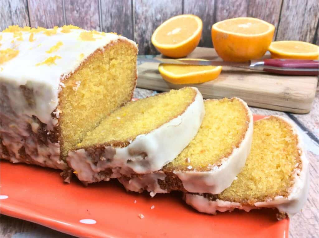 Sliced orange cake with white icing on an orange plate. Halved oranges and a red-handled knife rest on a wooden cutting board in the background—truly the best Orange Creamsicle Cake Recipe brought to life.