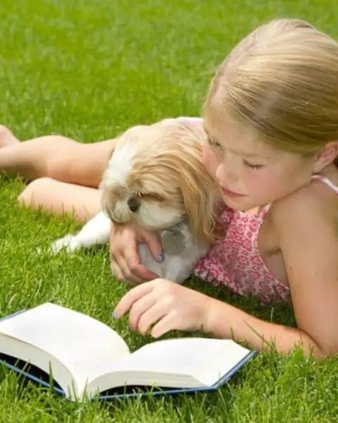 A little girl that is sitting in the grass with a book and a puppy.