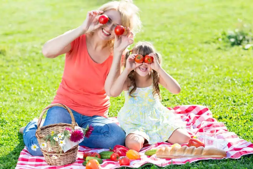 A mother and daughter holding produce up to their eyes and enjoying a summer time picnic.