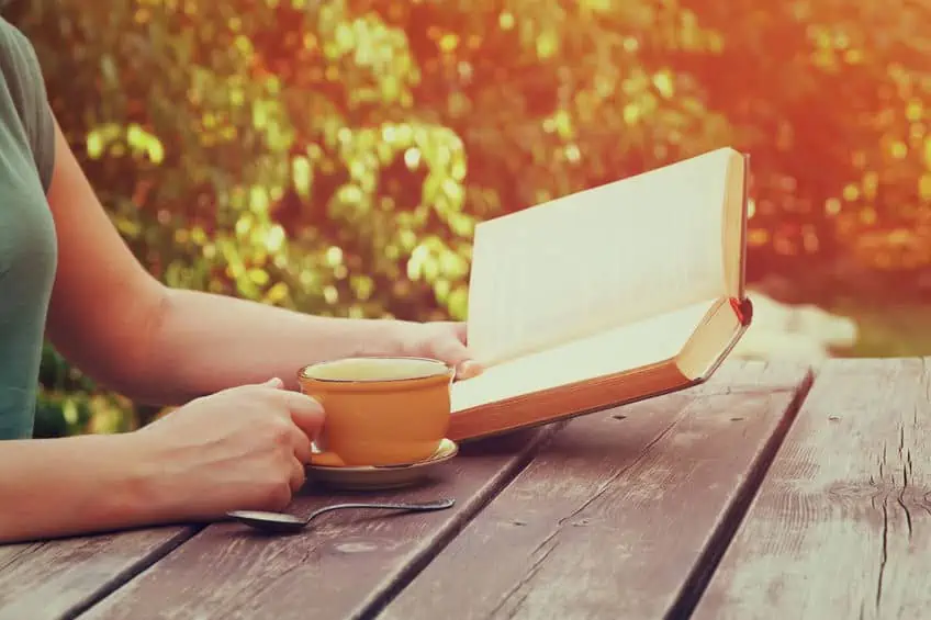 A person sitting on a bench in front of a picnic table reading a book and drinking a cup of coffee.