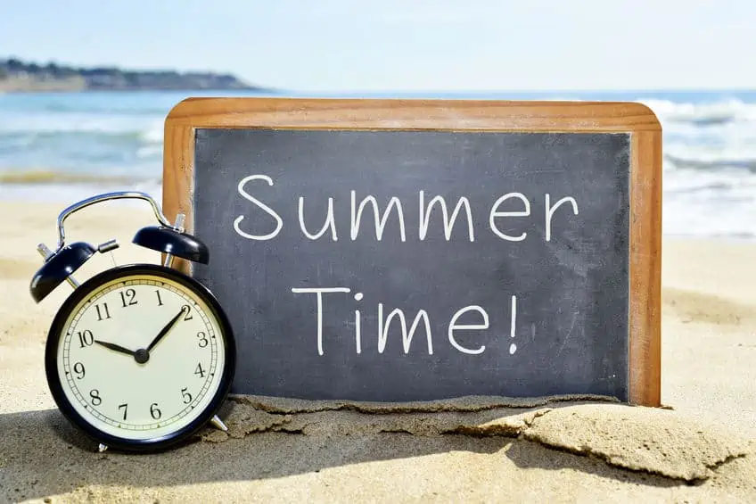 A clock sitting in front of a sign at the beach that reads \"summer time\".