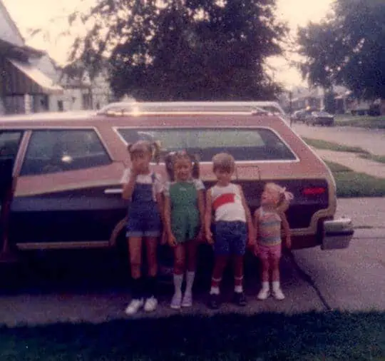 A family of children standing in front of an old car.