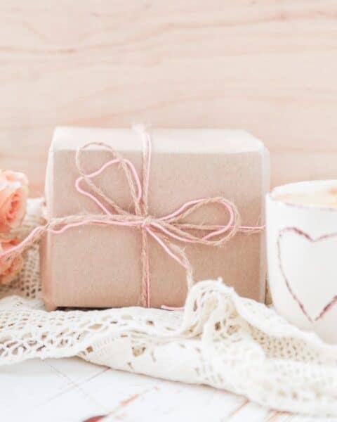 A wrapped package with a pink bow next to pink roses and a white cup on a lace tablecloth.