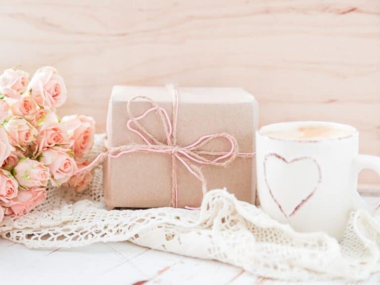 A wrapped package with a pink bow next to pink roses and a white cup on a lace tablecloth.