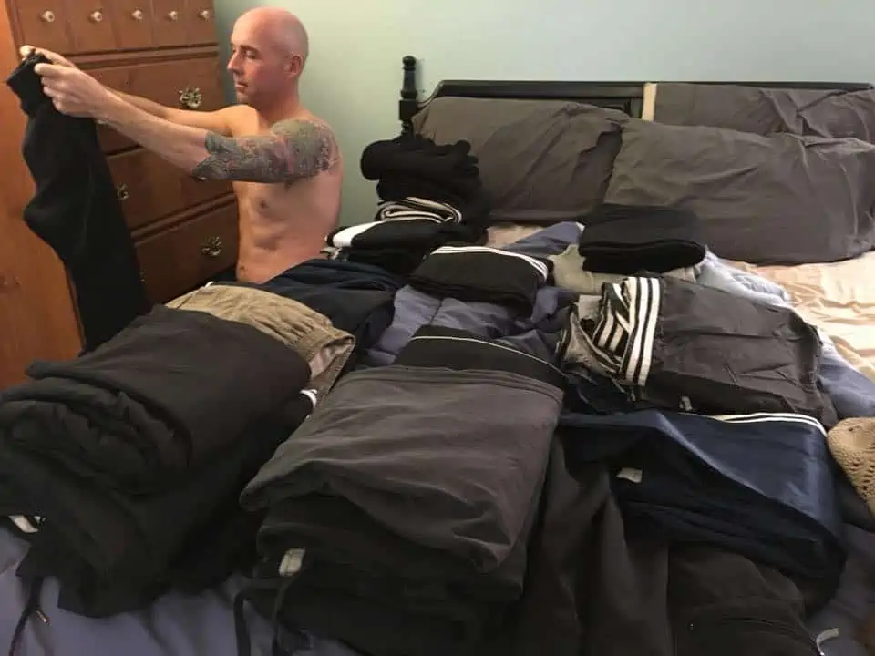 A man sitting on a bed with piles of clothes on the bed.