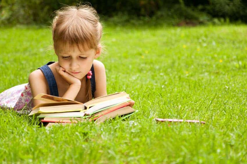 Child reading a stack of books in the grass.