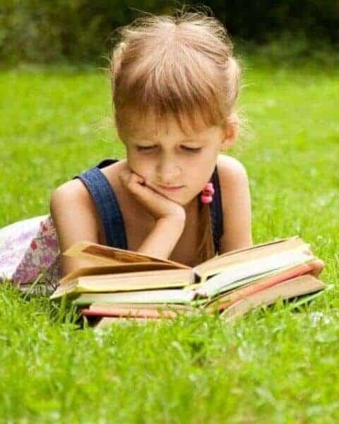 Child reading a stack of books in the grass.