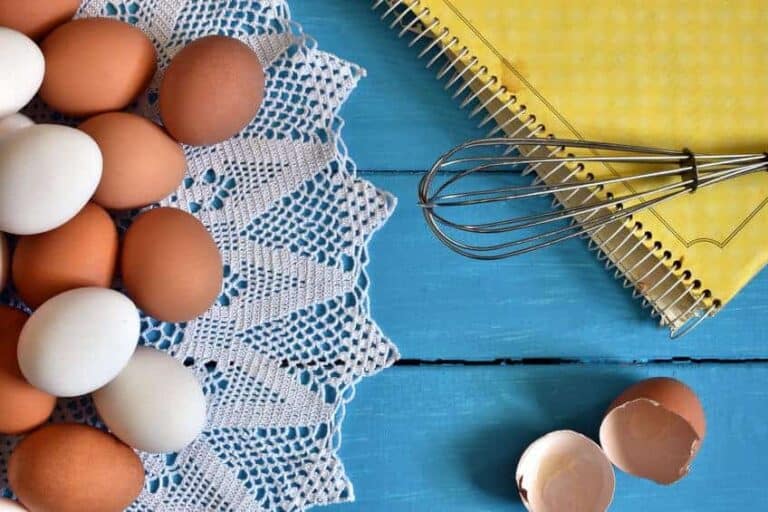 A group of eggs on a kitchen table with a whisk and cookbook.