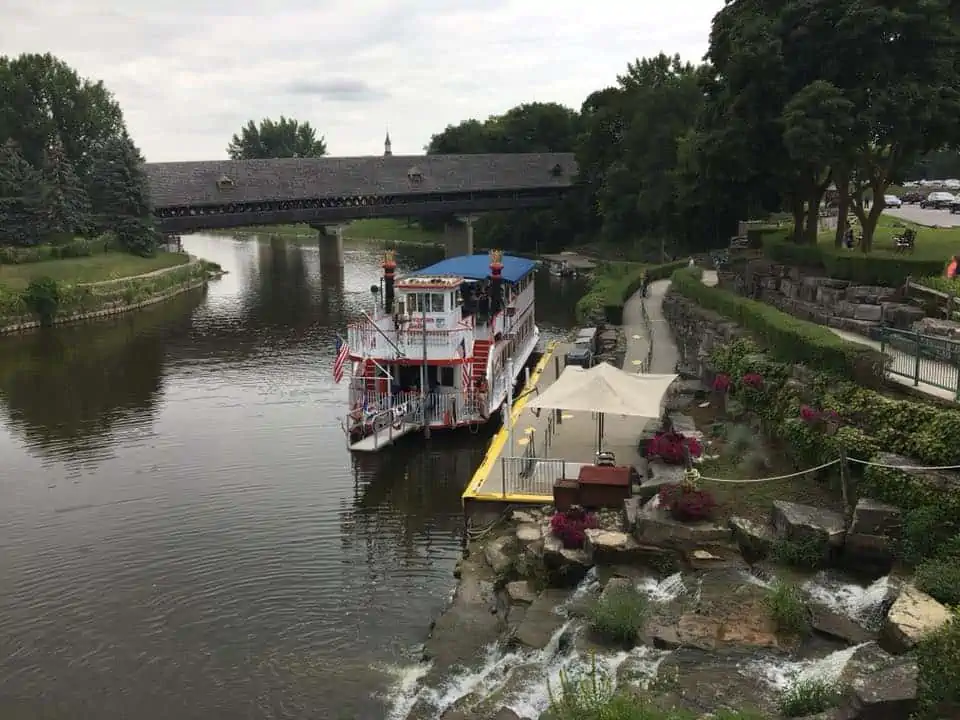 A boat traveling along a river next to a body of water. The Bavarian Inn Summertime Edition