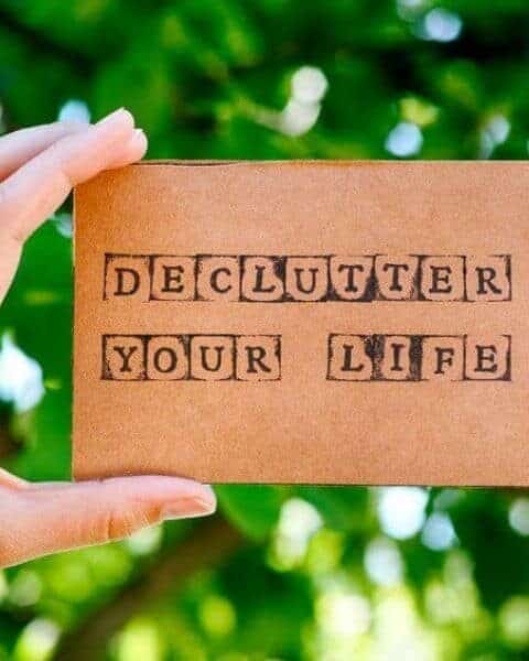 a womans hand holding a cardboard sign that says Declutter Your Life against a green leafy bush