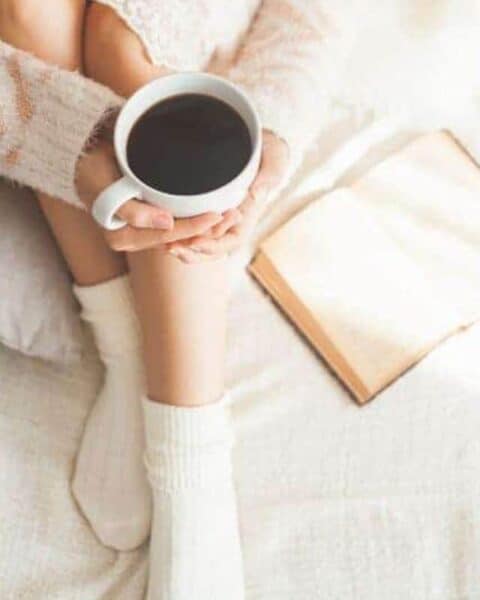 A woman holding a cup of coffee while sitting on her bed with a book open.