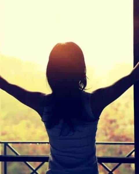 woman stading at a window holding open the drapes whiole the morning sun shines into the room
