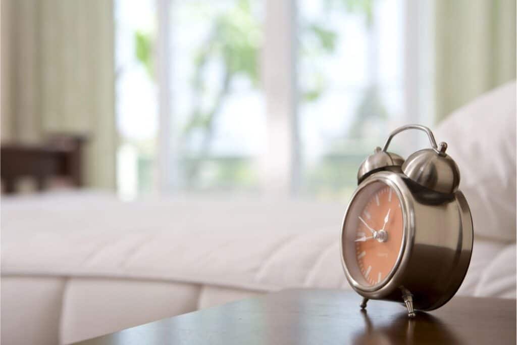 A metal analog alarm clock sits on a bedside table in a softly lit bedroom, helping with finding time in busy mornings, with a bed and large window in the background.