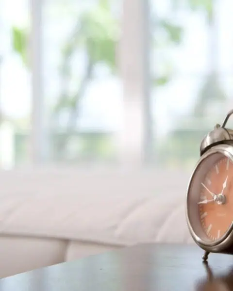 A metal analog alarm clock sits on a bedside table in a softly lit bedroom, helping with finding time in busy mornings, with a bed and large window in the background.