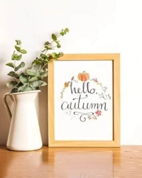 hello automn framed sign sitting ona wooden table next to a pitcher of flowers