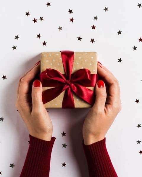 womans hands holding a wrapped Christmas gift with glitter sprinkled around it.