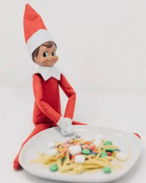 An elf on the shelf sitting next to a white plate filled with silly foods.