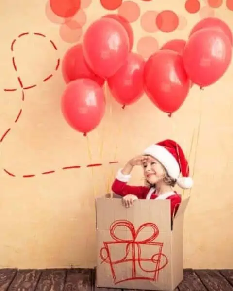 A little girl dressed as Santa Claus sitting in a cardboard box that's being lifted by balloons.