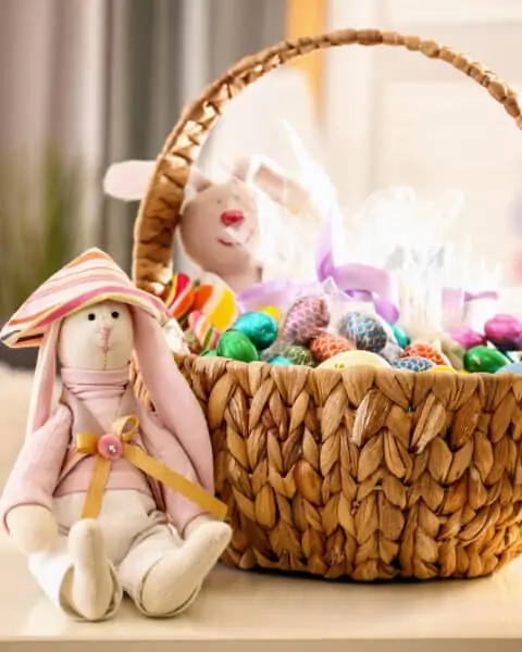 Beautiful Easter basket with traditional decorations and sweets near cute rabbit toy on table in light room