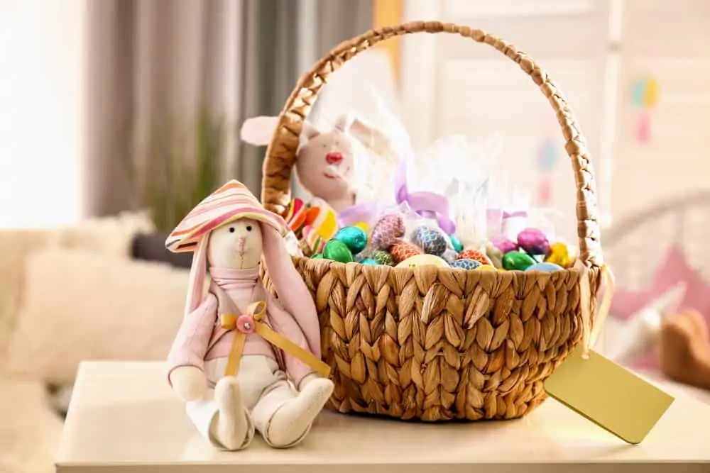 Beautiful Easter basket with traditional decorations and sweets near cute rabbit toy on table in light room
