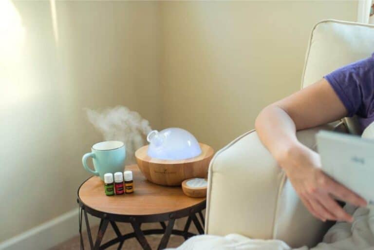 A person relaxes on the couch reading, while beside them, a small wooden table holds a mug and three aromatic bottles of essential oils. The essential oil diffuser emits mist, creating a soothing atmosphere, perhaps inspired by favorite essential oil diffuser recipes.